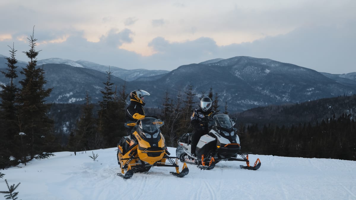 Two snowmobilers on a trail, with mountains in the background.