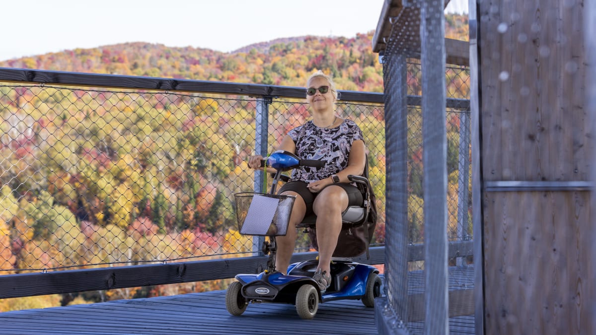 Femme en quadriporteur sur une passerelle en bois du Sentier des cimes, devant un paysage coloré.