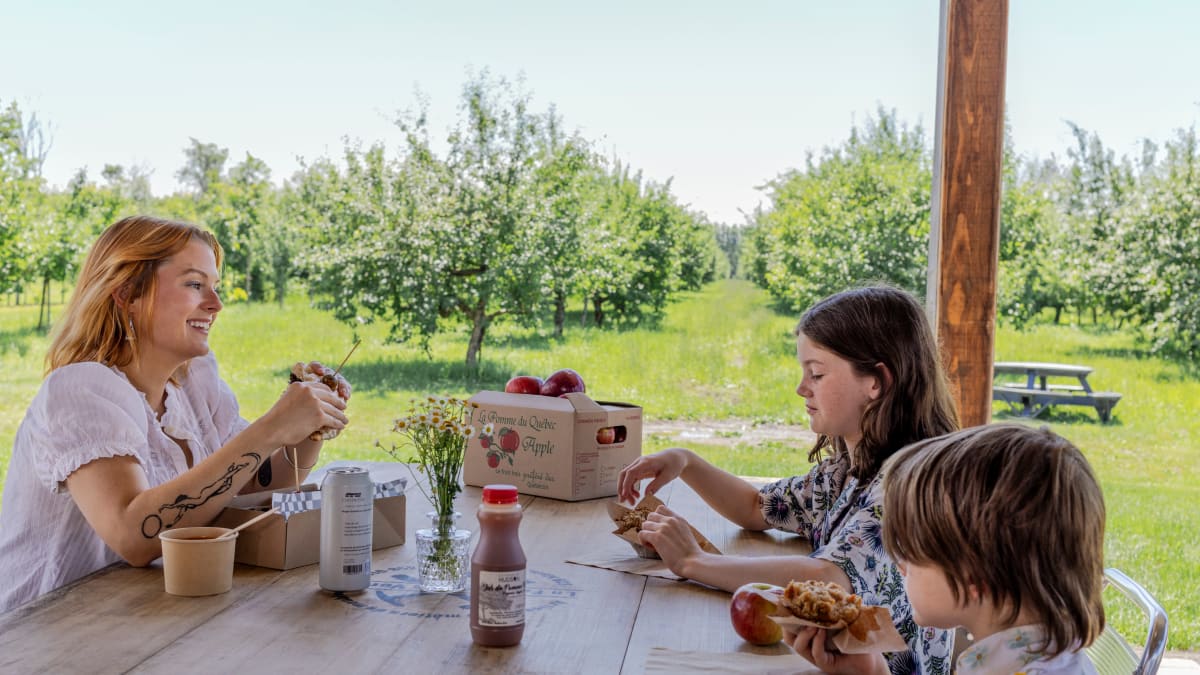 Une mère et ses deux enfants en train de déguster des produits de la pomme dans un verger.
