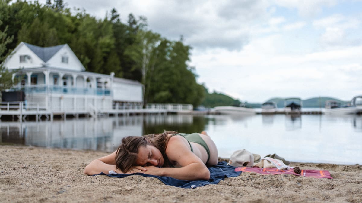 Femme étendue sur une plage; en arrière-plan, un plan d'eau et une auberge au bord de l'eau.