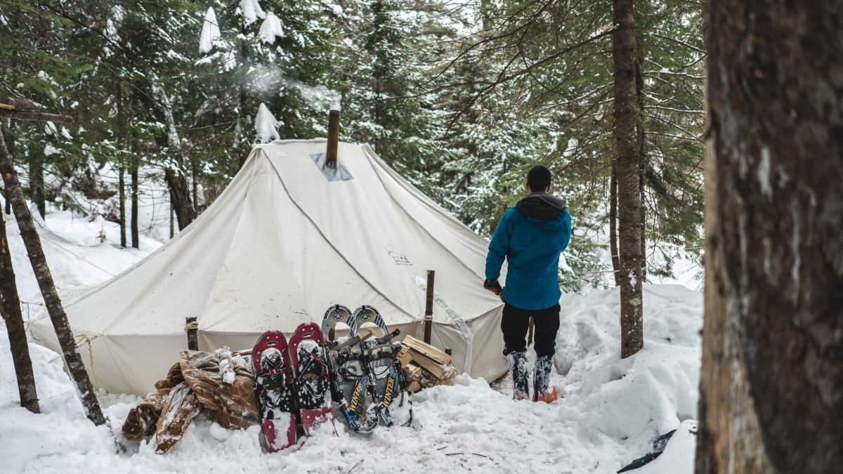 Personne devant une tente de camping hivernal, avec deux paires de raquettes plantées dans la neige à ses côtés.
