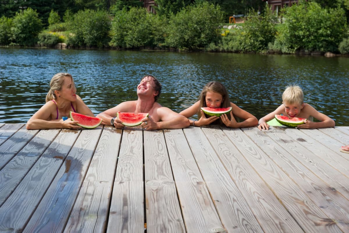 Four swimmers in the water, their arms resting on a dock, eating watermelon.