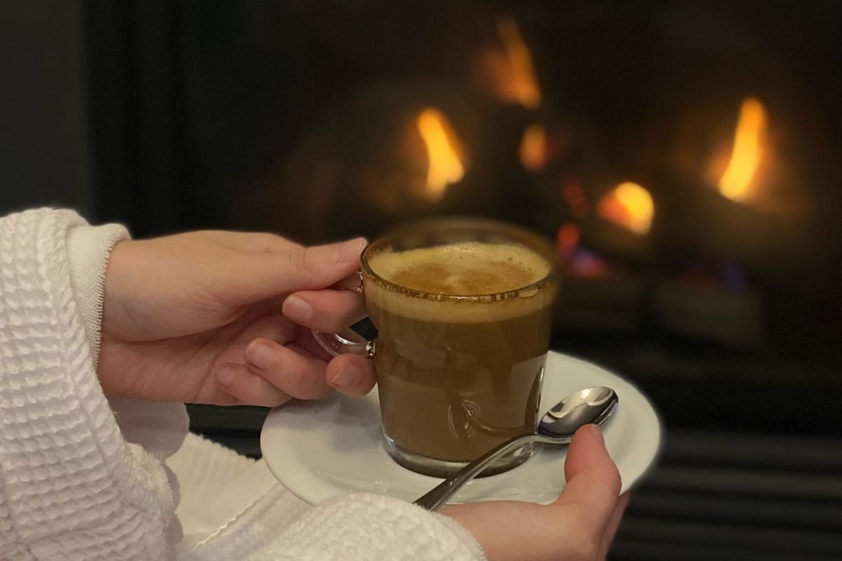 Woman in a bathrobe holding a coffee in front of the fireplace.