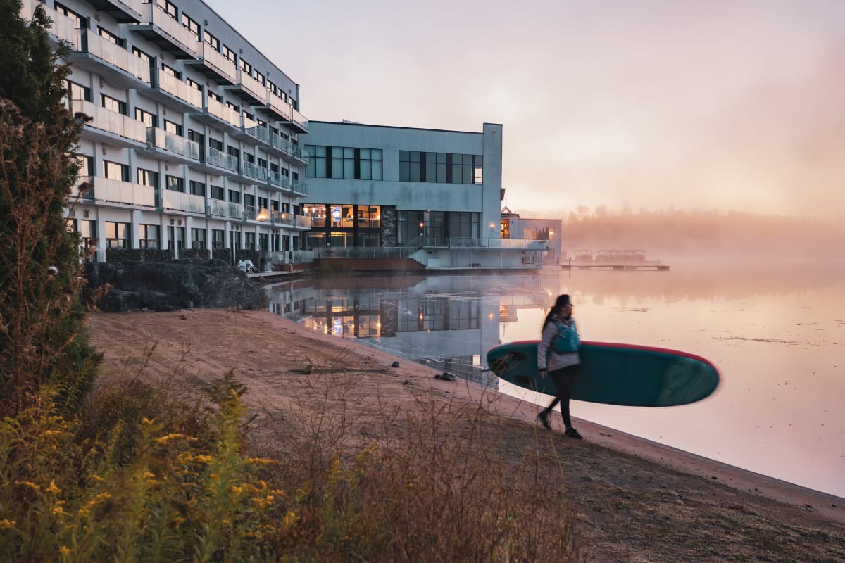 A woman holding a paddleboard at the edge of a lake at dawn.