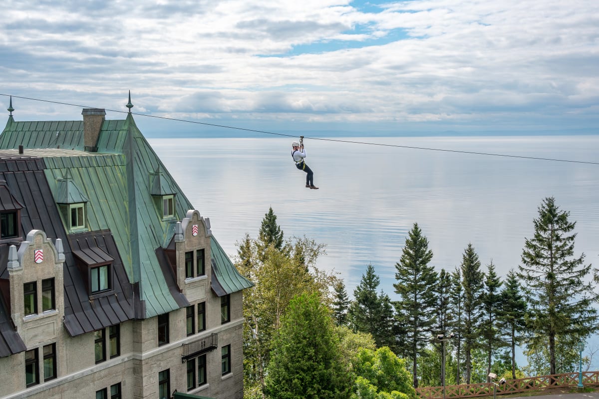 A person on a zipline above the Fairmont Le Manoir Richelieu, with the St. Lawrence River in the background.