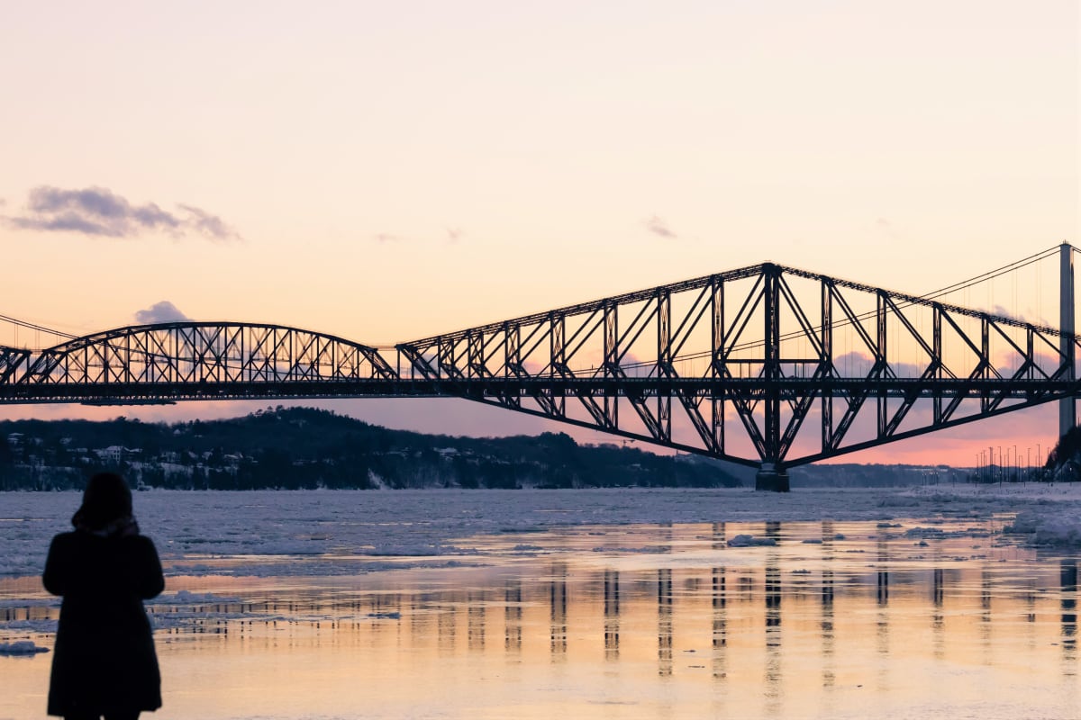 A person by the St. Lawrence River at sunset, with the Pont de Québec bridge on the horizon.