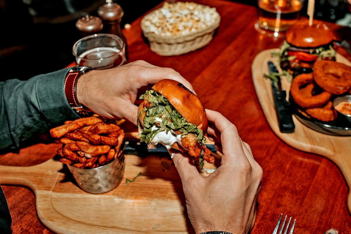 Two hands holding a hamburger above a board with fries on a restaurant table.