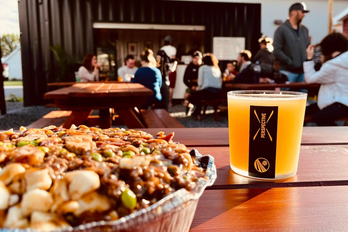 Gros plan sur une poutine et une bière; à l'arrière, des gens profitent de la terrasse de la microbrasserie Perspective.