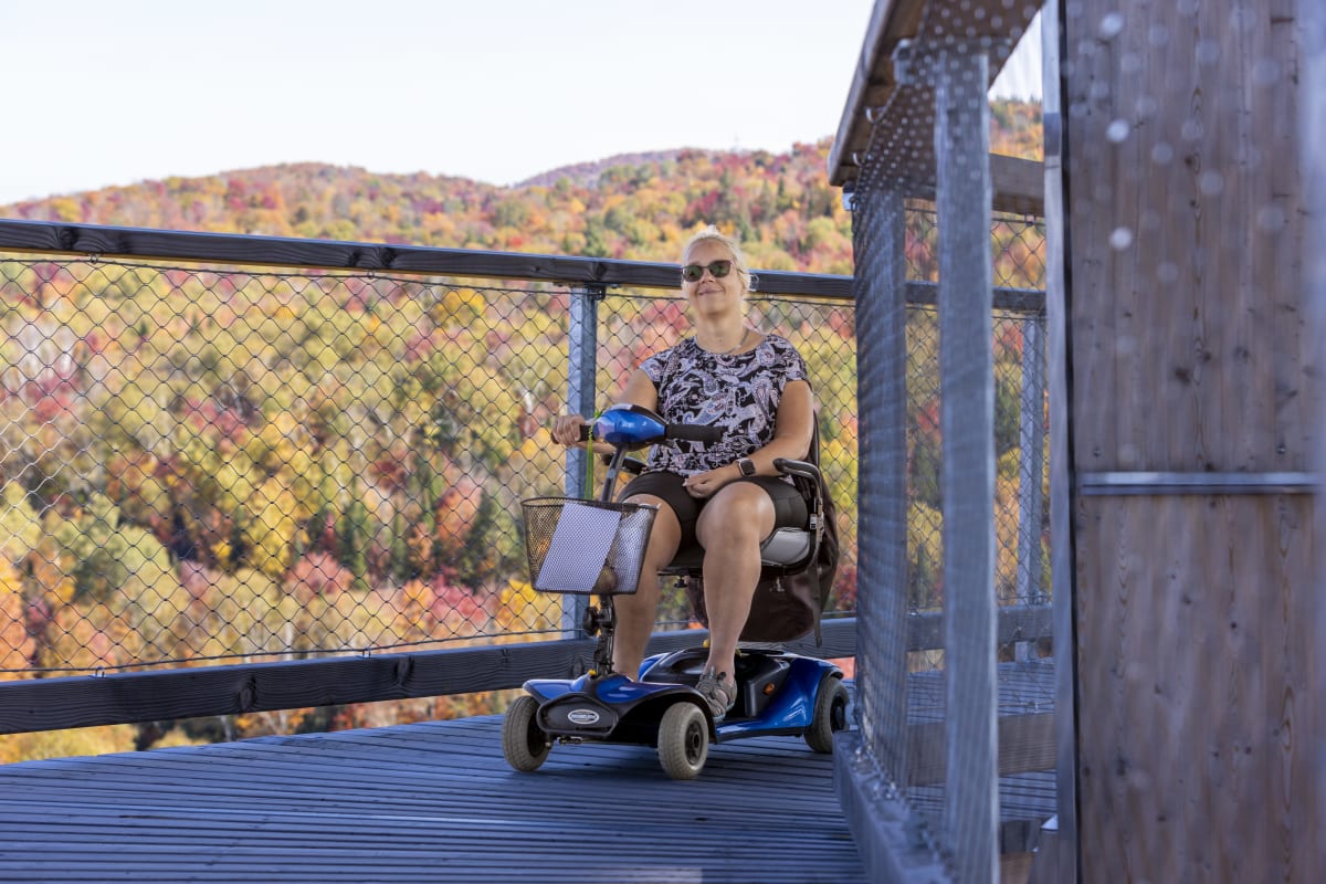 Femme en quadriporteur sur une passerelle en bois du Sentier des cimes, devant un paysage coloré.
