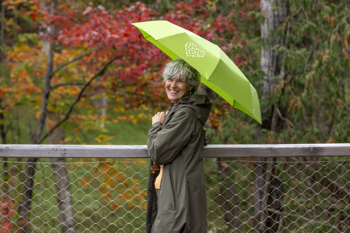 Femme souriante qui s'abrite sous un parapluie vert, avec un paysage automnal en arrière-plan.