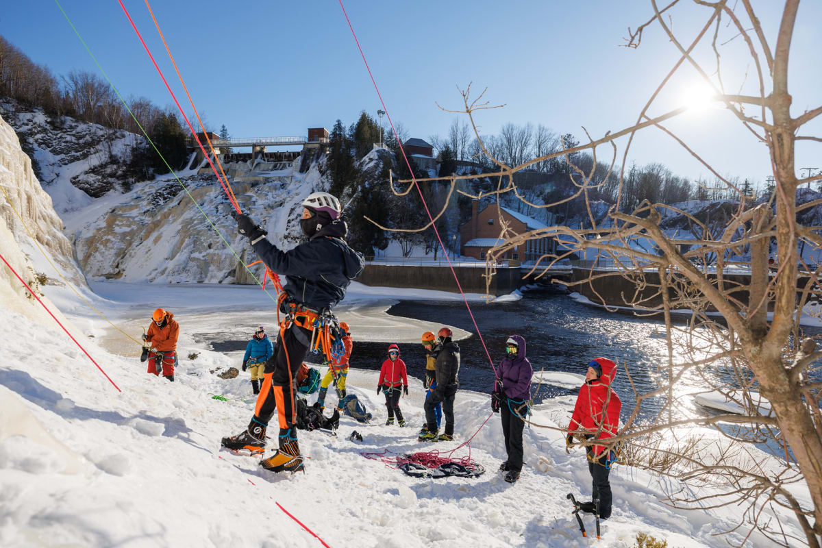 A person is about to start climbing an icy wall on a sunny day; behind them is a group of spectators.