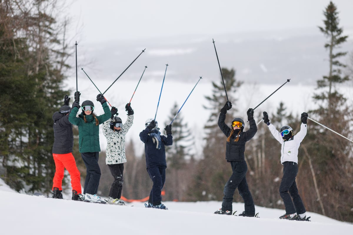 Group of six downhill skiers on a ski slope, arms raised in celebration. 