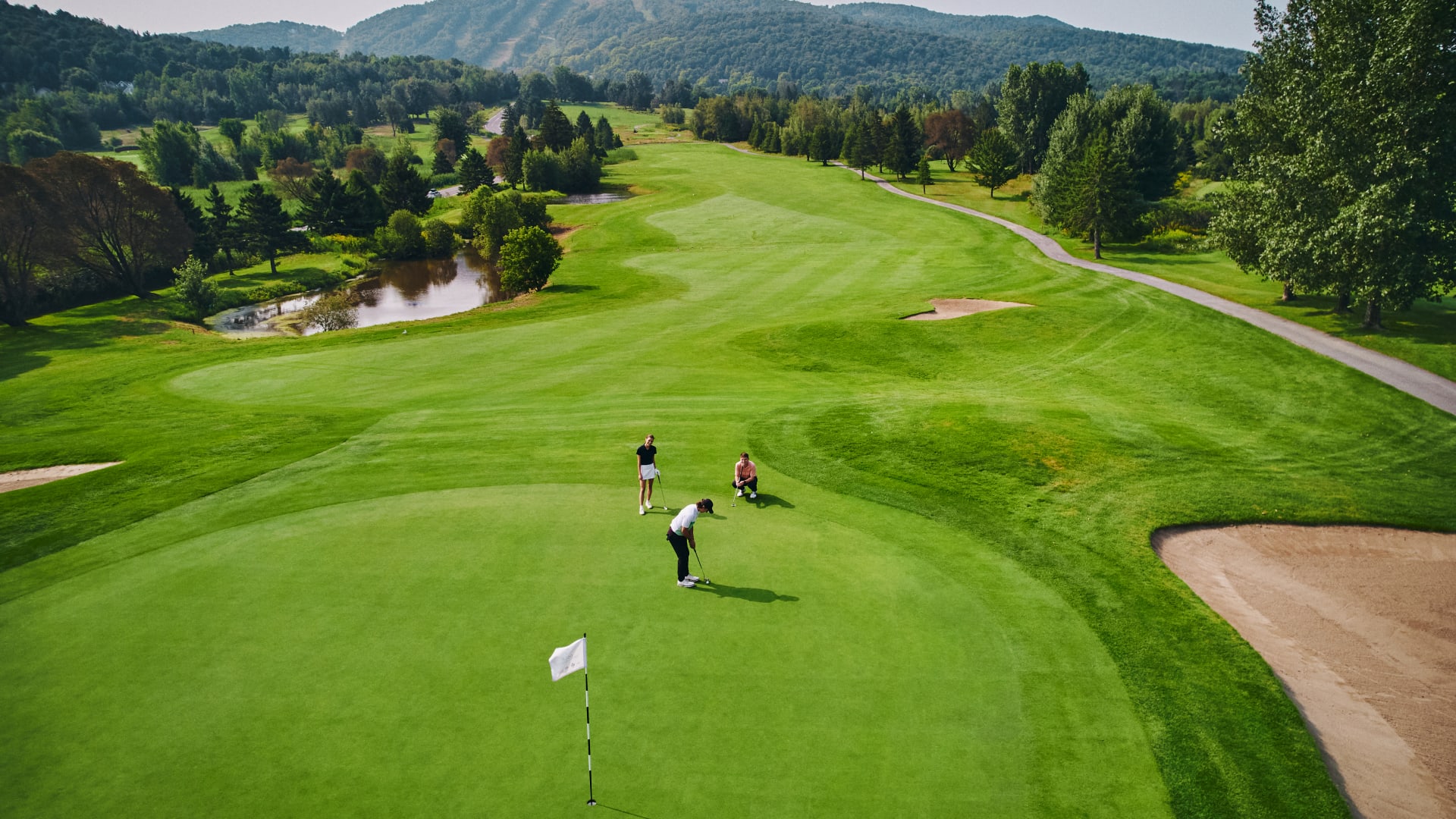 Trois personnes sur un vert au Golf Château-Bromont, avec la montagne en arrière-plan.