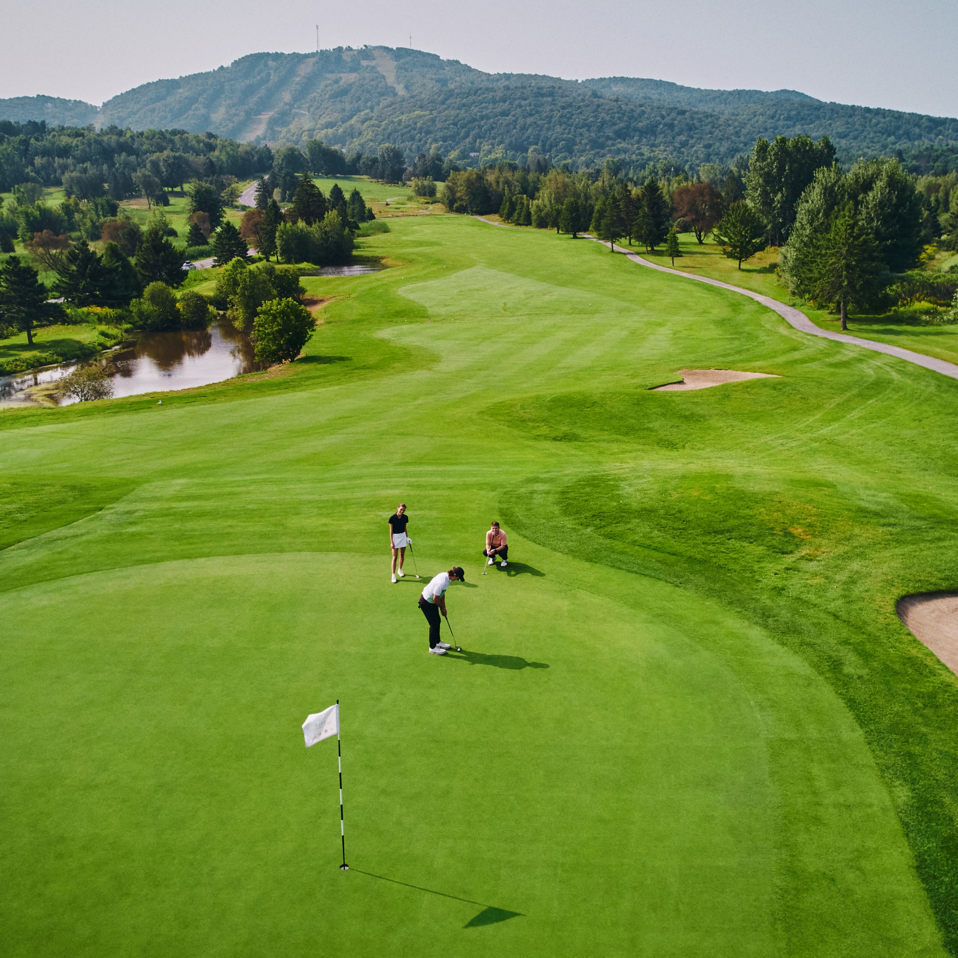 Trois personnes sur un vert au Golf Château-Bromont, avec la montagne en arrière-plan.
