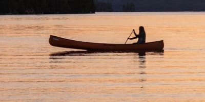 Une personne est assise dans un canot sur l'eau, avec un coucher de soleil en arrière-plan