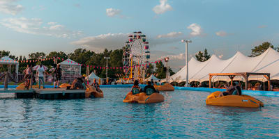 Festival Parasol - People on pedal boats in a large outdoor pool, with a Ferris wheel and white tents in the background.