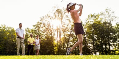 A woman plays golf, with three other individuals observing her in the background.