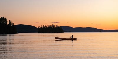 Une personne est assise dans un canot sur l'eau, avec un coucher de soleil en arrière-plan