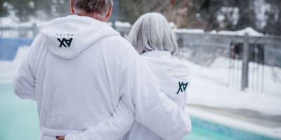 A couple in bathrobes embracing in front of a heated pool surrounded by snow.