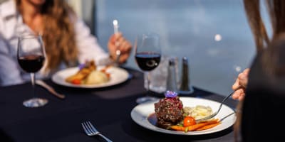 Two people enjoying a gourmet meal on an elegant table with a black tablecloth, including red wine and beautifully plated dishes, with a view of the water in the background.