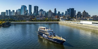 A cruise ship sailing on a river, with a panoramic view of a city skyline featuring tall skyscrapers in the background under a clear sky.