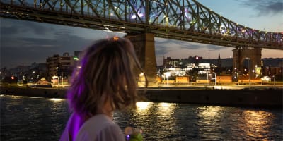 A woman viewed from behind holding a drink, admiring a lit-up bridge spanning a river, with city lights and a twilight sky in the background.