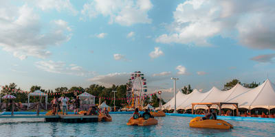 Festival Parasol - People on pedal boats in a large outdoor pool, with a Ferris wheel and white tents in the background.