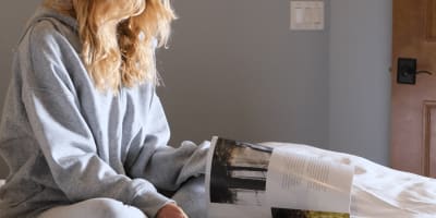 A young woman sitting on a bed, reading a magazine while enjoying her coffee.