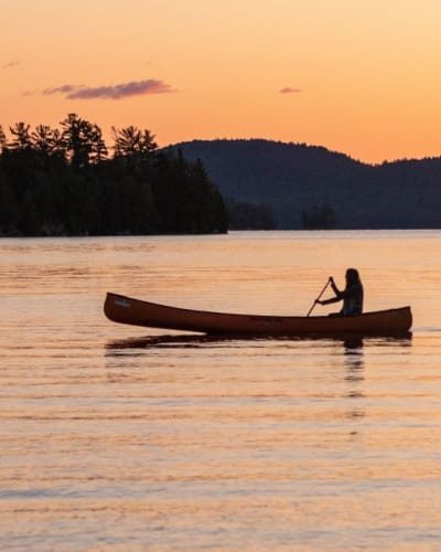 Une personne est assise dans un canot sur l'eau, avec un coucher de soleil en arrière-plan