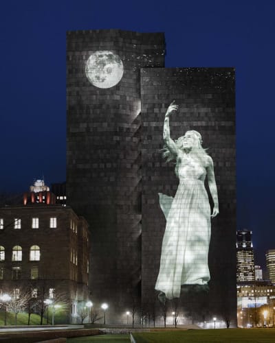 Cité Mémoire - Projection lumineuse d'une statue féminine et d'une lune sur un grand bâtiment sombre, la nuit