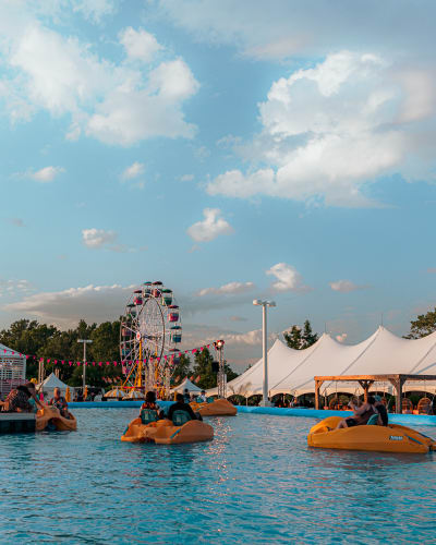 Festival Parasol - People on pedal boats in a large outdoor pool, with a Ferris wheel and white tents in the background.