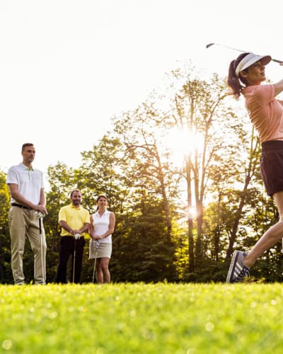 A woman plays golf, with three other individuals observing her in the background.