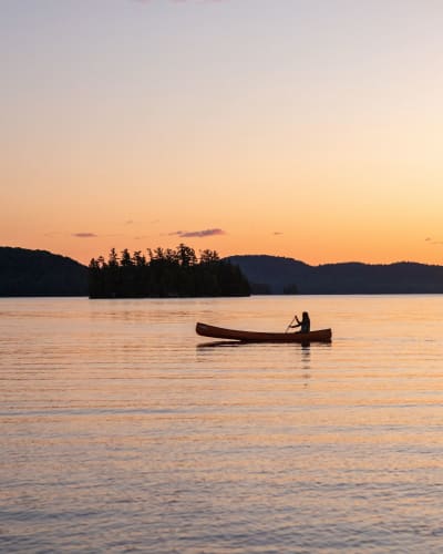 Une personne est assise dans un canot sur l'eau, avec un coucher de soleil en arrière-plan
