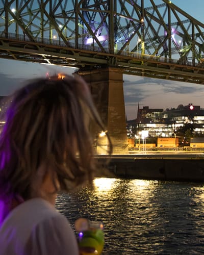 A woman viewed from behind holding a drink, admiring a lit-up bridge spanning a river, with city lights and a twilight sky in the background.