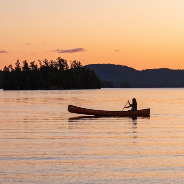 Une personne est assise dans un canot sur l'eau, avec un coucher de soleil en arrière-plan