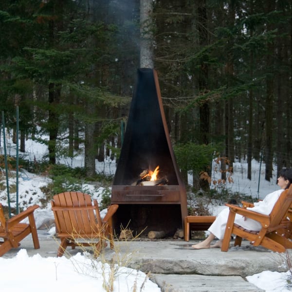 Femme assise sur une chaise adirondack à côté d'un foyer au spa à l'extérieur l'hiver.