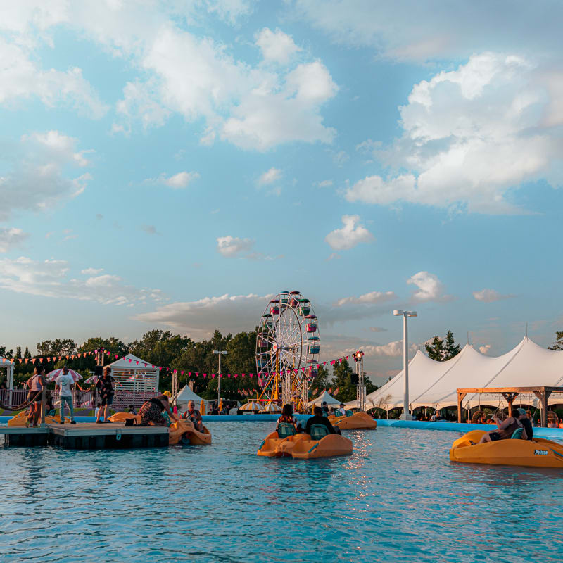 Festival Parasol - People on pedal boats in a large outdoor pool, with a Ferris wheel and white tents in the background.