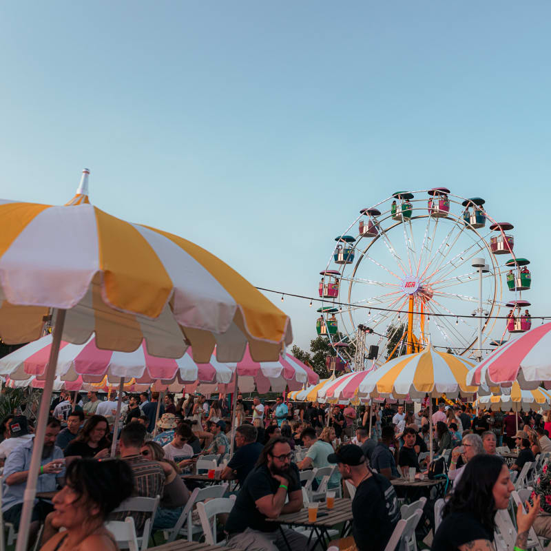 Festival Parasol - People are sitting at tables with umbrellas, with a Ferris wheel in the background.