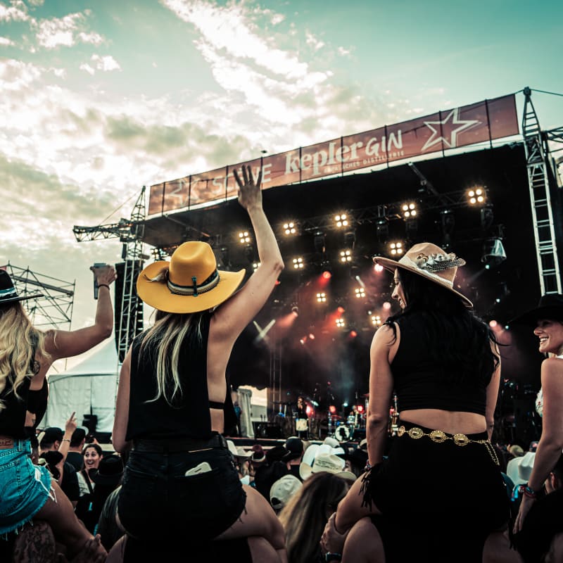 Crowd in front of an outdoor stage including four women wearing cowboy hats.