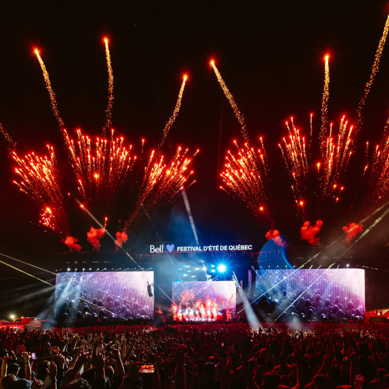 Crowd in front of an outdoor stage at night with fireworks.