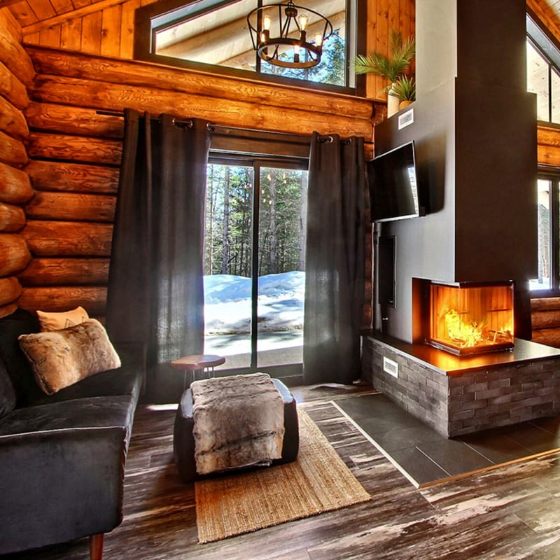 Interior of a log cabin with a central fireplace and large windows.