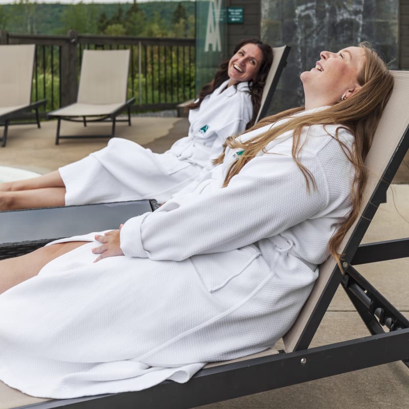 Two women in bathrobes laughing while lying on lounge chairs.