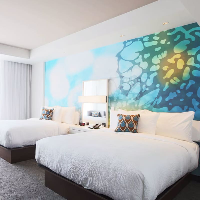 Chambre d’hôtel illuminée de lumière naturelle, avec deux grands lits, literie blanche et mur d’accent bleu.