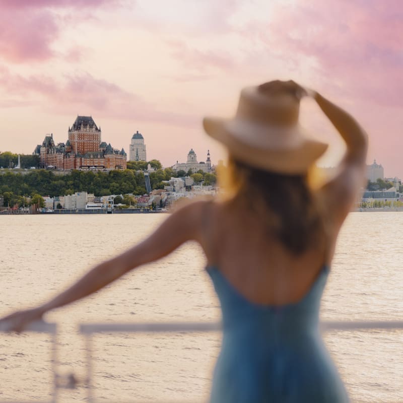 A woman in a blue dress and a large hat looks out over the Château Frontenac from the St. Lawrence River.