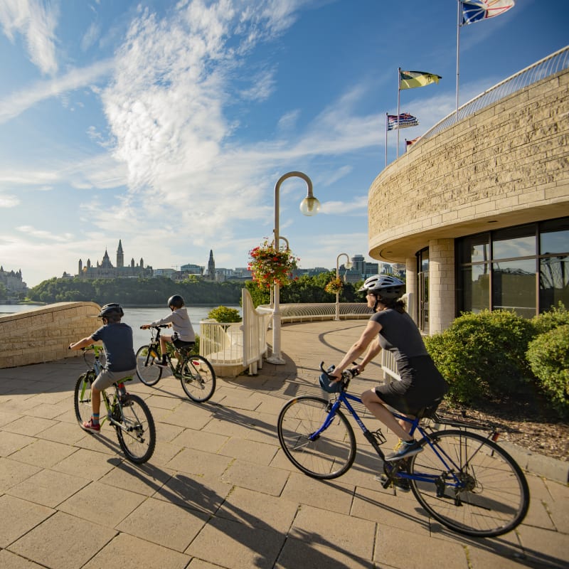 Mother and two sons on bikes next to the Canadian Museum of History in front of the Ottawa River.