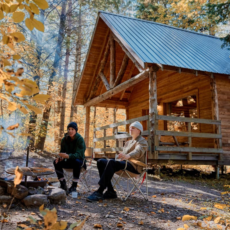 Two people sitting around a campfire in front of a wooden cottage in a forest in autumn.