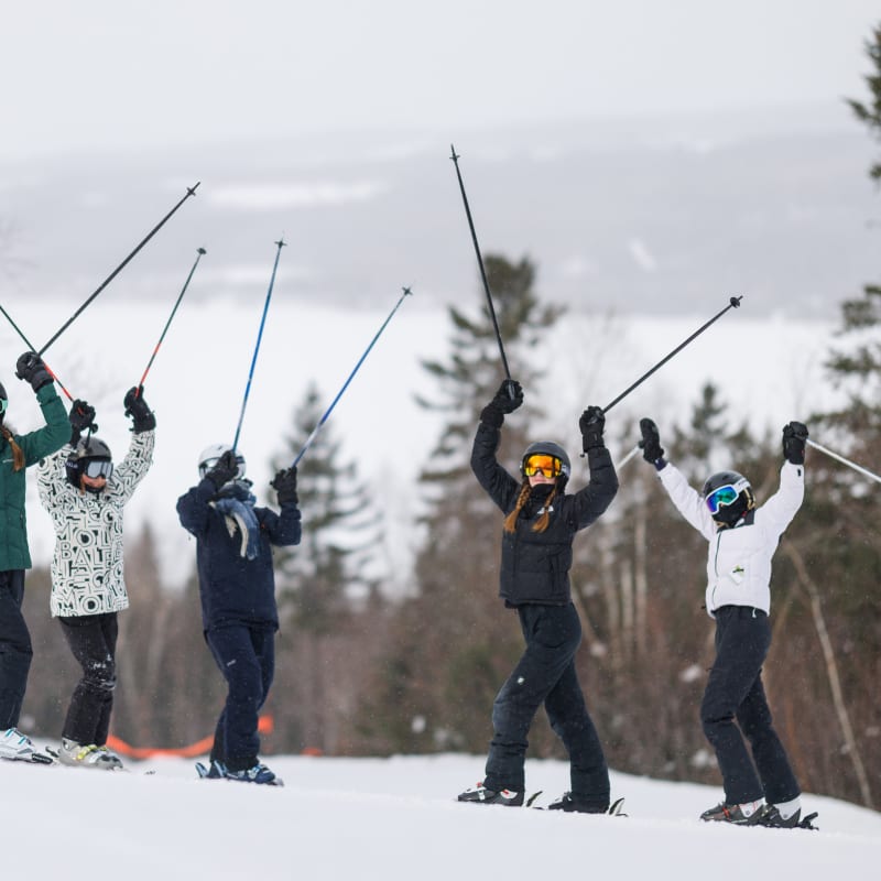 6 skiers at the top of the mountain proudly lifting their poles in the air.