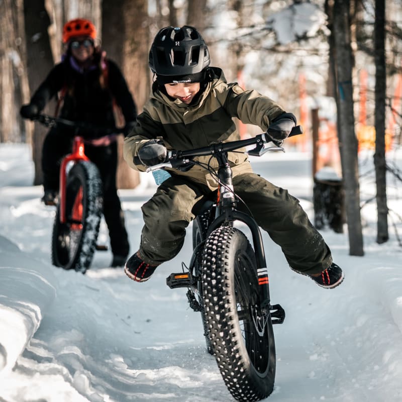 Jeune garçon en fatbike dans une forêt enneigée en avant-plan, avec une autre personne plus loin derrière.