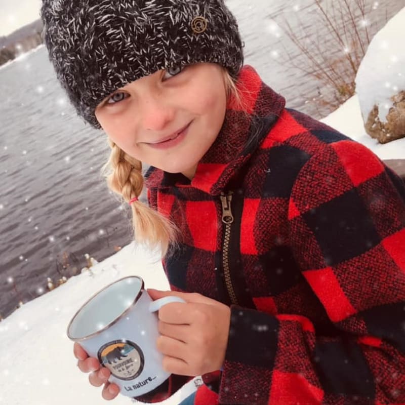 Young girl with a hat and plaid shirt by the water in the winter holding a mug