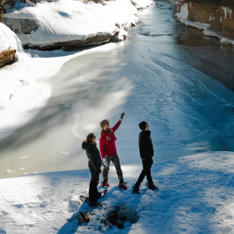 3 people wearing snowshoes crossing a river in the forest in winter.